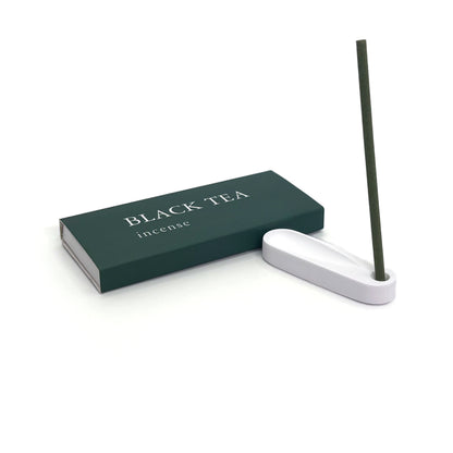 Burning black tea incense stick placed in minimalist plaster holder next to green box