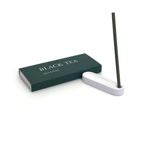Burning black tea incense stick placed in minimalist plaster holder next to green box